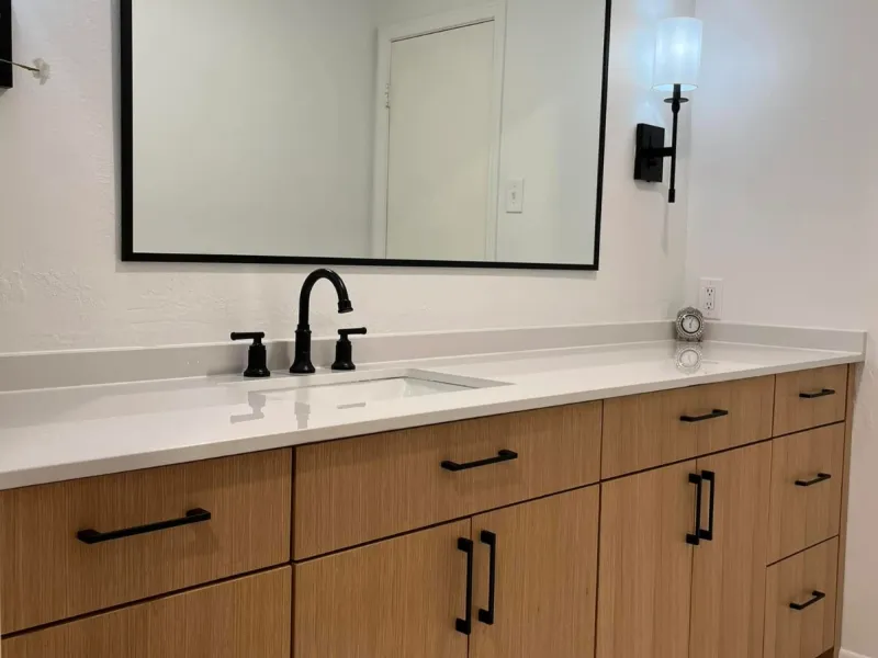 Long light-oak bathroom vanity with a white counter, black faucet, black-framed mirror, and herringbone floor tile.