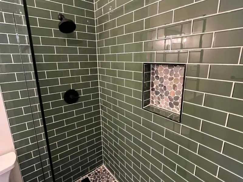 Shower lined in green subway tile with a river-pebble floor, matching pebble niche, black fixtures, and a glass panel.